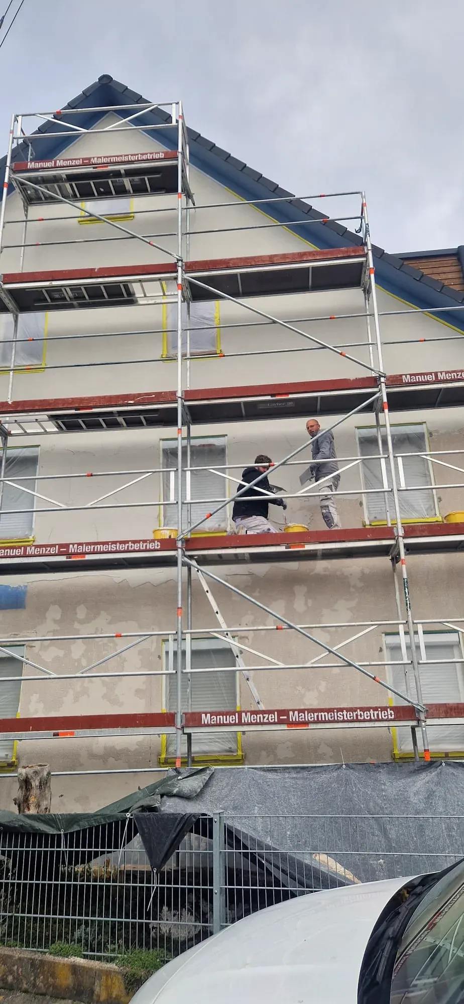 Zwei Maler von Malermeister Menzel spachteln auf dem Gerüst die Fassade eines Wohnhauses, an den Gerüstböden der Schriftzug „Manuel Menzel – Malermeisterbetrieb“